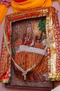 A framed artwork depicting a religious or mythological scene. At the center, a deity-like figure with a halo and ornate attire sits serenely. Below, a figure appears to be lying on a fire, possibly in a sacrificial or ritualistic manner. The background includes a swastika, trees, a horse, and additional human figures. The frame is elaborately decorated with red and gold fabric, floral designs, and a string of pearls.