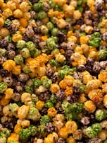 A close-up view of multicolored popcorn, featuring vibrant shades of green, brown, yellow, and white. Each piece is coated with assorted flavors and appears crunchy and appetizing.