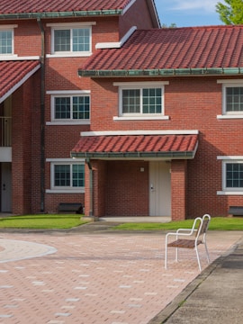 A brick building with a red tiled roof and multiple white-framed windows. There is a small covered porch area with a white door. In the foreground, a single bench with metal armrests is placed on a paved surface. The surrounding area is neatly landscaped with green grass.