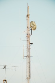 A tall communication tower with metal lattice structure and multiple antennas, including a large dish antenna, all set against a clear blue sky.