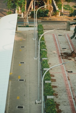 An aerial view of a paved area featuring two parallel paths lined with greenery. Street lamps are positioned along the paths, and the area appears to be well-maintained with a few fallen leaves scattered. Trees and a gated entrance can be seen in the background.