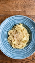 A plate of creamy pasta topped with shredded cheese is presented on a blue dish. The pasta appears to be fettuccine alfredo, with a rich and smooth sauce. The table beneath has a wooden texture, adding warmth to the setting.