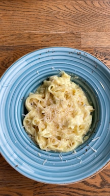 A plate of creamy pasta topped with shredded cheese is presented on a blue dish. The pasta appears to be fettuccine alfredo, with a rich and smooth sauce. The table beneath has a wooden texture, adding warmth to the setting.