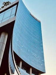 A modern, curved skyscraper with glass windows reflects the light. The building has a sleek, contemporary design with a large logo near the top. The sky is clear, adding a sense of openness and height.