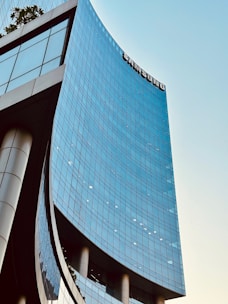 A modern, curved skyscraper with glass windows reflects the light. The building has a sleek, contemporary design with a large logo near the top. The sky is clear, adding a sense of openness and height.
