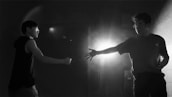Two people face each other in a shadowy, dimly lit space. One person extends their hand while the other appears to be in a defensive posture. A bright light source behind creates dramatic contrasts and shadows.
