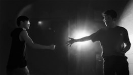 Two people face each other in a shadowy, dimly lit space. One person extends their hand while the other appears to be in a defensive posture. A bright light source behind creates dramatic contrasts and shadows.
