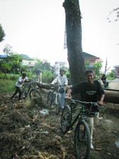 A group of young men is seen pushing mountain bikes on a dirt path through a wooded area. One of them is smiling, and they appear to be enjoying an outdoor adventure. The environment is lush with greenery, and there is a mix of trees and houses in the background.