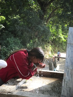 A person wearing a red jacket is drinking water with cupped hands from a stone fountain outdoors, surrounded by dense greenery. There is another person walking in the background on a dirt path.