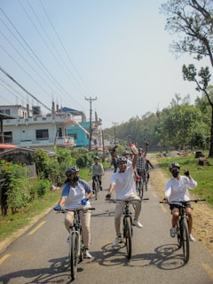 A group of people are riding bicycles on a road lined with trees and houses. They appear to be enjoying the ride, wearing helmets and casual attire. Some are gesturing excitedly, creating a lively atmosphere.