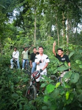 A group of five young men are standing with their bicycles in a dense, green forest. They appear to be enjoying a break during a cycling trip, surrounded by lush foliage and tall trees. Some of them are gesturing with their hands, adding a lively and adventurous vibe to the scene.