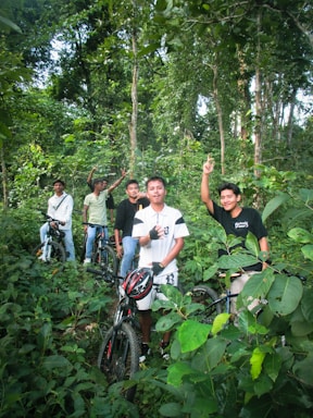A group of five young men are standing with their bicycles in a dense, green forest. They appear to be enjoying a break during a cycling trip, surrounded by lush foliage and tall trees. Some of them are gesturing with their hands, adding a lively and adventurous vibe to the scene.