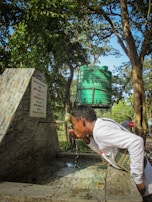 A person is drinking water from a faucet in an outdoor setting. The scene includes a stone structure with an inscription and a green water tank in the background, surrounded by trees and greenery.