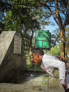 A person is drinking water from a faucet in an outdoor setting. The scene includes a stone structure with an inscription and a green water tank in the background, surrounded by trees and greenery.