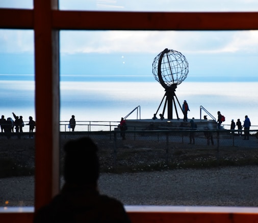 A silhouetted view through a window highlights a group of people gathered around a large globe sculpture on a coastal promontory. The background features a serene, expansive seascape under a mostly clear sky.