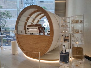 A modern, stylish coffee shop display featuring a large, cylindrical coffee counter with a wooden and white design. The counter has an espresso machine with the % Arabica logo. To the right are transparent shelves holding various branded merchandise, including bags, cups, and bottles. The setting is in a clean, polished building interior with glass walls showing an urban street view.