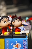 Two cartoon character figurines with large eyes and cheerful expressions are sitting on display, surrounded by various colorful toys and objects. One of the figurines is depicted on a blue box with Japanese text, featuring the character from the Doraemon series.