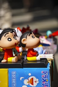 Two cartoon character figurines with large eyes and cheerful expressions are sitting on display, surrounded by various colorful toys and objects. One of the figurines is depicted on a blue box with Japanese text, featuring the character from the Doraemon series.
