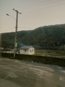 A rural landscape with a small white house surrounded by a natural setting. In the background, there is a densely wooded hill, and a road with power lines and a streetlight runs across the foreground. The scene appears calm and undisturbed, with muted colors contributing to a serene atmosphere.