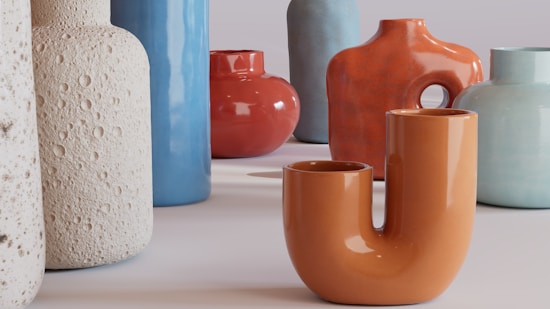 A collection of ceramic vases with varied shapes and textures displayed on a surface. The vases exhibit different colors including orange, blue, and beige, with some having smooth surfaces while others have a more textured appearance.