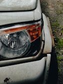 Close-up view of a car's headlight and front corner. The white paint is worn and rusty around the edges, indicating age and exposure to the elements. The headlight is intact and features an orange indicator on its side. The surface of the car is slightly dirty, with the ground visible in the background.