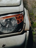Close-up view of a car's headlight and front corner. The white paint is worn and rusty around the edges, indicating age and exposure to the elements. The headlight is intact and features an orange indicator on its side. The surface of the car is slightly dirty, with the ground visible in the background.
