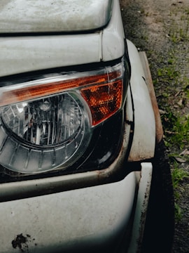 Close-up view of a car's headlight and front corner. The white paint is worn and rusty around the edges, indicating age and exposure to the elements. The headlight is intact and features an orange indicator on its side. The surface of the car is slightly dirty, with the ground visible in the background.