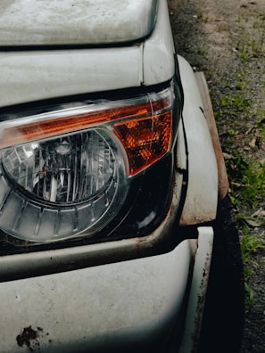 Close-up view of a car's headlight and front corner. The white paint is worn and rusty around the edges, indicating age and exposure to the elements. The headlight is intact and features an orange indicator on its side. The surface of the car is slightly dirty, with the ground visible in the background.
