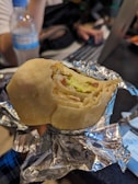 A partially eaten wrap or burrito held by a hand, wrapped in aluminum foil. The contents include vegetables such as lettuce and possibly some type of meat. In the background, there is a plastic water bottle on a table and a person using a laptop.