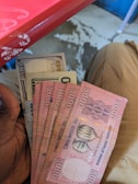 Several banknotes held in a hand, including Rwandan 5000 franc notes and a US 100-dollar bill. The background shows part of a red table and a person wearing beige pants seated.