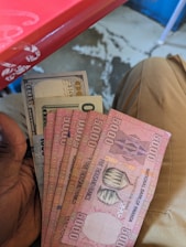 Several banknotes held in a hand, including Rwandan 5000 franc notes and a US 100-dollar bill. The background shows part of a red table and a person wearing beige pants seated.