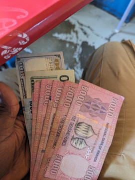 Several banknotes held in a hand, including Rwandan 5000 franc notes and a US 100-dollar bill. The background shows part of a red table and a person wearing beige pants seated.