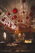 A sophisticated dining area adorned with numerous hanging red ornaments. The ceiling features an intricate leaf pattern, adding texture and elegance. Warm, ambient lighting creates a cozy and inviting atmosphere. Tables are set with plates and glasses, ready for diners.
