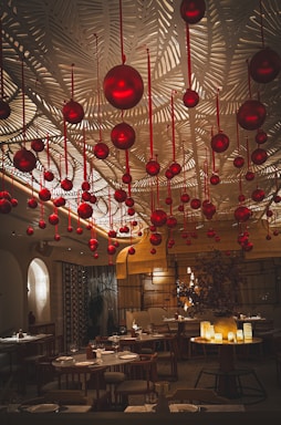 A sophisticated dining area adorned with numerous hanging red ornaments. The ceiling features an intricate leaf pattern, adding texture and elegance. Warm, ambient lighting creates a cozy and inviting atmosphere. Tables are set with plates and glasses, ready for diners.