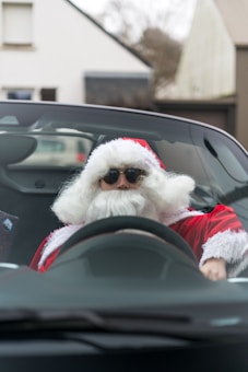 A person dressed in a Santa Claus outfit, complete with a red suit and white beard, is sitting in the driver's seat of a convertible car, wearing sunglasses. The background shows a suburban setting with houses and a blurred car.