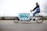 A person is riding a cargo bike with a large compartment that has a colorful design featuring the word 'pony'. The rider is dressed casually in a blue vest, jeans, and white sneakers, traveling on a paved path. In the background, there is a metal fence and some leafless trees, suggesting a cool or overcast day.