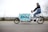 A person is riding a cargo bike with a large compartment that has a colorful design featuring the word 'pony'. The rider is dressed casually in a blue vest, jeans, and white sneakers, traveling on a paved path. In the background, there is a metal fence and some leafless trees, suggesting a cool or overcast day.