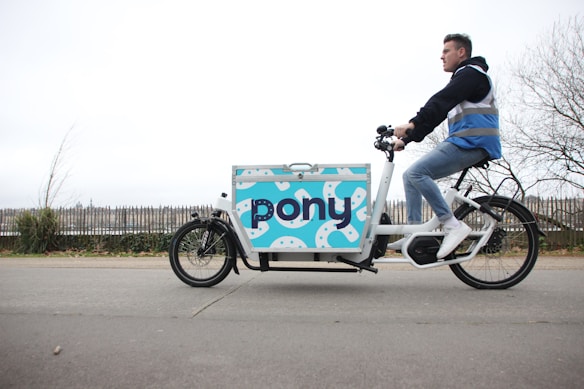 A person is riding a cargo bike with a large compartment that has a colorful design featuring the word 'pony'. The rider is dressed casually in a blue vest, jeans, and white sneakers, traveling on a paved path. In the background, there is a metal fence and some leafless trees, suggesting a cool or overcast day.