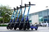 A row of e-scooters is parked in an outdoor area near a modern building with large glass windows. The scooters are aligned neatly, each with a blue and purple color scheme. Trees in the background add a touch of greenery to the urban environment.