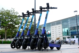A row of e-scooters is parked in an outdoor area near a modern building with large glass windows. The scooters are aligned neatly, each with a blue and purple color scheme. Trees in the background add a touch of greenery to the urban environment.