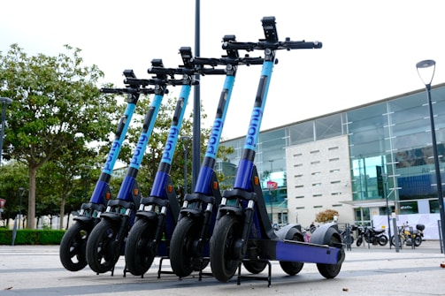 A row of e-scooters is parked in an outdoor area near a modern building with large glass windows. The scooters are aligned neatly, each with a blue and purple color scheme. Trees in the background add a touch of greenery to the urban environment.
