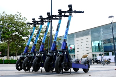 A row of e-scooters is parked in an outdoor area near a modern building with large glass windows. The scooters are aligned neatly, each with a blue and purple color scheme. Trees in the background add a touch of greenery to the urban environment.