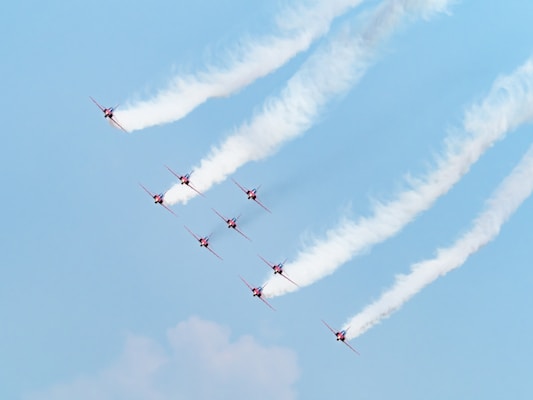 A formation of eight red jets flying in the sky, leaving white smoke trails behind them. The planes are aligned in a V-shape pattern with blue sky in the background.