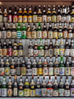 A collection of various bottles, mostly sake, arranged neatly in rows. The bottles have colorful labels with Japanese characters and illustrations, showcasing a variety of designs and brands. The arrangement creates a visually appealing pattern.