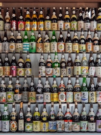 A collection of various bottles, mostly sake, arranged neatly in rows. The bottles have colorful labels with Japanese characters and illustrations, showcasing a variety of designs and brands. The arrangement creates a visually appealing pattern.