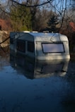 A caravan is partially submerged in water surrounded by a flooded environment. The scene is set outdoors with trees in the background and reflections visible in the calm water.