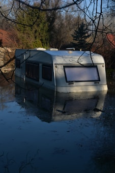 A caravan is partially submerged in water surrounded by a flooded environment. The scene is set outdoors with trees in the background and reflections visible in the calm water.