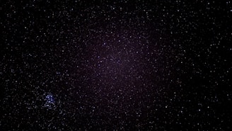 A starry night sky filled with countless tiny specks of light spread across a dark canvas. In the bottom left, a cluster of brighter stars stands out, forming a distinct grouping.