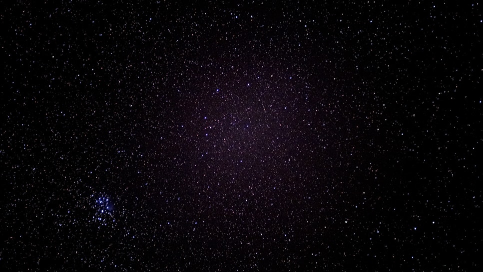 A starry night sky filled with countless tiny specks of light spread across a dark canvas. In the bottom left, a cluster of brighter stars stands out, forming a distinct grouping.