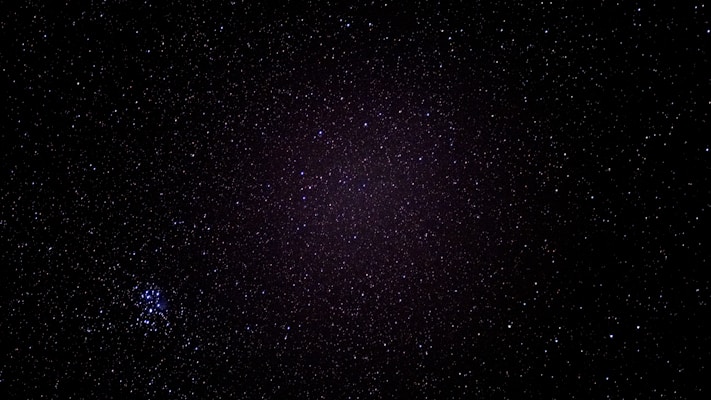 A starry night sky filled with countless tiny specks of light spread across a dark canvas. In the bottom left, a cluster of brighter stars stands out, forming a distinct grouping.